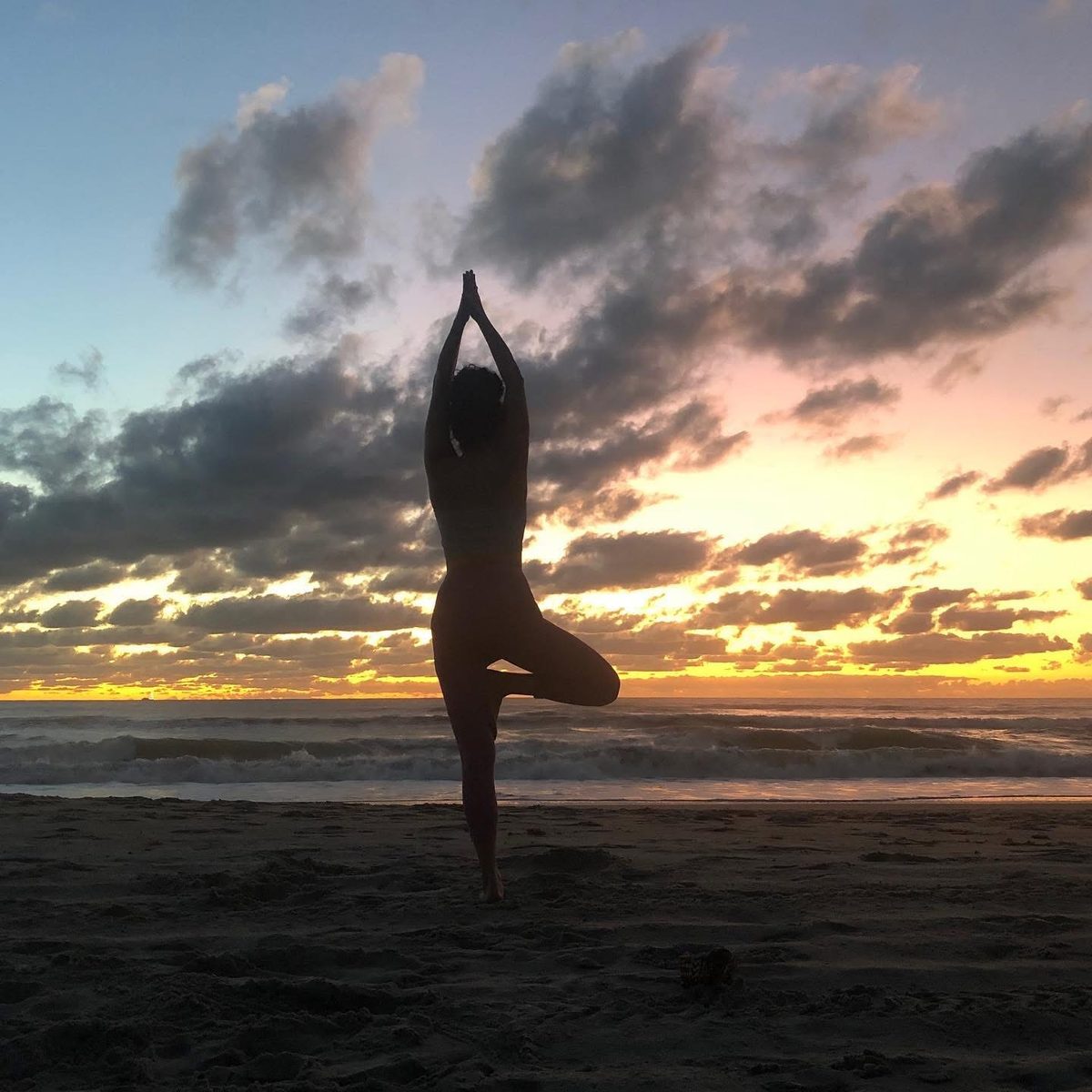 Silhouette of a woman in tree pose against a sunset over the ocean