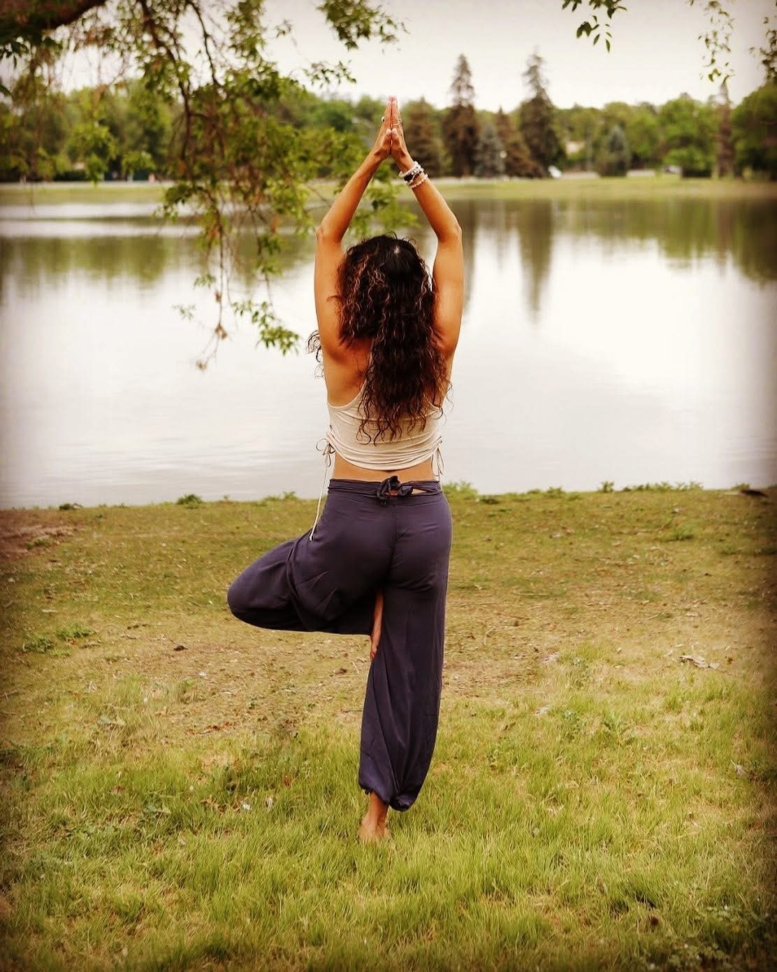 Woman in tree pose facing a lake, arms raised in prayer position, practicing yoga outdoors